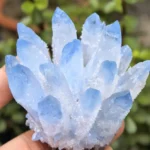 A close-up of a hand holding a raw aquamarine phantom crystal cluster outdoors, showcasing its icy blue points against a soft green garden background