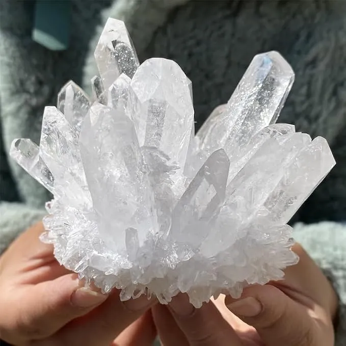 A close-up of hands holding a raw clear quartz crystal cluster, showcasing its pristine icy points against a soft, textured gray background