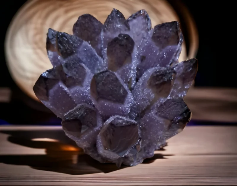 A natural ametrine phantom crystal cluster displayed under dramatic lighting on a slatted wooden surface, highlighting its deep purple and grey hues.