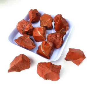 Several natural raw red jasper crystal chunks displayed on and around a small, light-colored square tray against a white surface, featuring earthy terracotta tones with subtle white quartz veining.