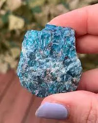 A close-up macro view of fingers holding a single raw blue apatite crystal, showcasing its rich blue color and reflective, highly textured natural surface.