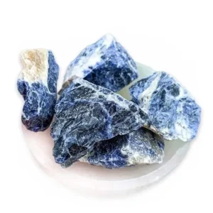 A close-up view of a small pile of raw sodalite crystal chunks resting on a translucent, shallow dish, highlighting their vibrant blue hues mixed with natural white and beige inclusions