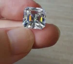 A loose Asscher cut moissanite stone held lightly between two fingers, displaying a mesmerizing "hall of mirrors" effect and flashes of rainbow fire within its step-cut facets.