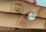 A sparkling loose Asscher cut moissanite stone resting between two fingers to demonstrate its physical size and clear step-cut facets.