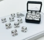 A collection of loose Asscher cut moissanite stones scattered on a white display pad, with six matching stones neatly arranged inside a small black gem display box.
