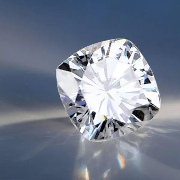 An artistic, dramatically lit macro shot of a cushion cut moissanite resting on a reflective surface against a soft blue background, displaying intense light return and internal reflections.