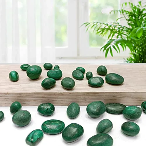 Lifestyle display of polished natural green Malachite tumbled stones arranged across a light wooden board and white tabletop beside a lush green indoor plant for Heart Chakra healing and Vastu energy balance.