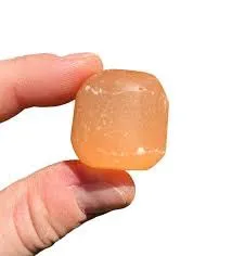 Macro close-up of a single barrel-polished natural Peach Orange Selenite tumbled stone held between thumb and forefinger against a pure white background, showcasing warm apricot translucency and silky satin-spar striations for Sacral Chakra healing.